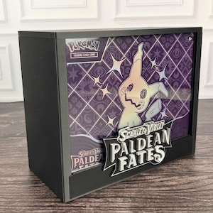 May include: A black display case containing a Pokemon Scarlet and Violet Paldean Fates trading card game box. The box features a purple diamond pattern background with a Mimikyu illustration and the text Scarlet and Violet Paldean Fates.