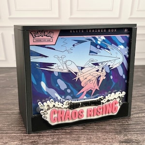 May include: A black display case containing a Pokemon Elite Trainer Box with a blue and pink graphic featuring a stylised bird creature. The front of the case features a white and pink layered sign with the text Chaos Rising in bold letters.