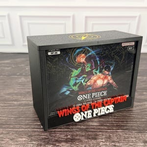 Könnte beinhalten: Ein schwarzer rechteckiger Schaukasten mit einer Booster-Box für das One Piece Wings of the Captain Kartenspiel. Die Box zeigt Illustrationen von zwei Charakteren, den Text Wings of the Captain, One Piece Card Game und 12 Karten pro Packung.