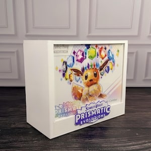 Può includere: Una scatola espositiva rettangolare bianca con una grafica Pokémon Scarlet and Violet Prismatic Evolutions. L'illustrazione mostra un Eevee circondato da cristalli fluttuanti colorati. Il testo Prismatic Evolutions è stampato sul pannello frontale.