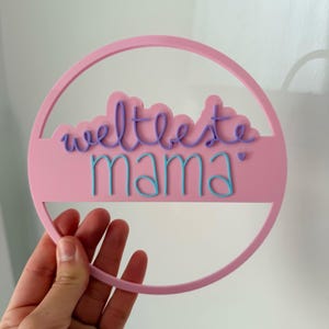 Könnte beinhalten: Ein rosa kreisförmiges Schild mit den Worten "weltbeste mama" in lila und blauer Schrift. Das Schild wird von einer Hand vor einem weißen Hintergrund gehalten. Das Schild hat einen kreisförmigen Rahmen.