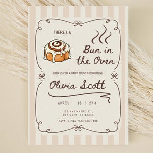 May include: A baby shower invitation with a tan and white striped background. The invitation features a cartoon cinnamon roll and the text "There's a Bun in the Oven." It also includes the name Olivia Scott, date, time, and address.