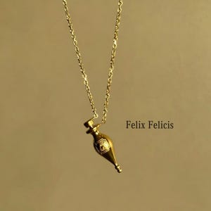Può includere: Una collana color oro con un ciondolo a forma di boccetta di pozione. La boccetta ha un piccolo emblema ed è sospesa a una delicata catena. Il testo "Felix Felicis" è visibile nell'immagine.