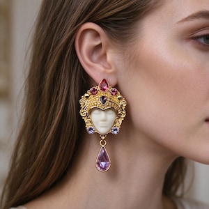 Puede incluir: Un pendiente llamativo en tono dorado con un camafeo central de rostro esculpido en blanco rodeado de metal ornamentado. El diseño está acentuado con cristales rosas, morados y rojo oscuro, e incluye un cristal rosa colgante en forma de lágrima.