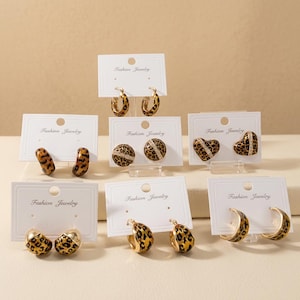 Może przedstawiać: Osiem par kolczyków prezentowanych na białych kartonikach z napisem Fashion Jewelry. Kolczyki wykonane są ze złotego metalu w brązowo-czarne cętki lamparta, a niektóre wzory zawierają małe przezroczyste kryształki i kształty serc.