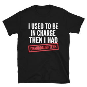 Può includere: T-shirt nera con testo bianco: "I USED TO BE IN CHARGE THEN I HAD GRANDDAUGHTERS". La parola "GRANDDAUGHTERS" è in un riquadro rosso. La t-shirt è a maniche corte e girocollo. Il testo è in grassetto e senza grazie.