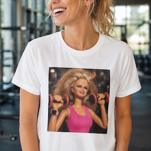 May include: White crew neck t-shirt with a graphic of a doll lifting weights. The doll has blonde hair, a pink top, and is holding red dumbbells. The doll is wearing a pink smartwatch.