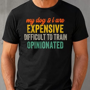 Puede incluir: Una camiseta negra de manga corta con el texto "my dog & i are EXPENSIVE DIFFICULT TO TRAIN OPINIONATED" impreso en letras de bloque de color naranja, amarillo, blanco y turquesa con una textura desgastada de estilo vintage en el pecho.