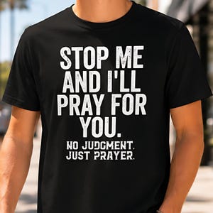 Op de afbeelding: Een zwart t-shirt met korte mouwen en witte tekst in een verweerde stijl die luidt: STOP ME AND I'LL PRAY FOR YOU. NO JUDGMENT. JUST PRAYER. Het shirt heeft een ronde hals en een standaard pasvorm, met de afbeelding gecentreerd op de borst.