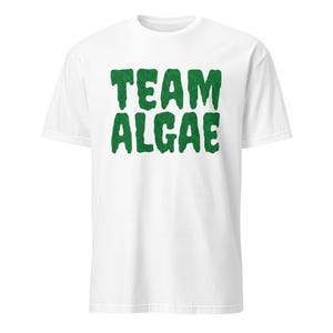 May include: A white short-sleeved t-shirt featuring the text "TEAM ALGAE" printed in a bold, dark green font with a textured, dripping, or slime-like appearance centered on the chest area of the garment.
