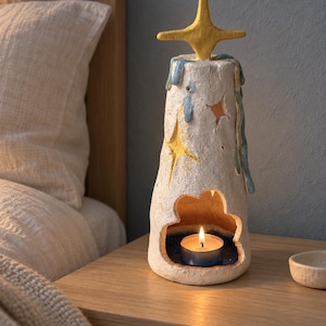 May include: A textured, off-white ceramic candle holder shaped like a tall, tapered tower. It features a yellow star topper, blue drip accents, and star-shaped cutouts. A cloud-shaped opening at the base contains a small, lit tea light candle.