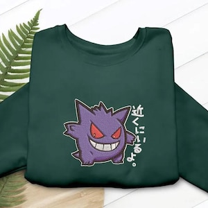 Peut inclure: Un sweat-shirt à col rond vert foncé avec une broderie représentant le personnage Gengar violet aux yeux rouges et au large sourire. À droite du personnage, le texte japonais "近くにいるよ。" est brodé en fil blanc sur la poitrine.