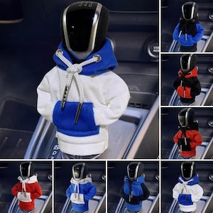 Puede incluir: Una funda para el pomo de la palanca de cambios del coche con forma de sudadera con capucha en miniatura. La sudadera es blanca con detalles azules en la capucha y el bolsillo. El cordón es blanco. También se muestran fundas adicionales en rojo, negro y azul.