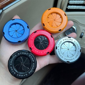May include: A collection of five car engine start/stop button covers in various colors: blue, orange, red, silver, and black. Each cover has the words "ENGINE START STOP" around the edge and a stylized design in the center.