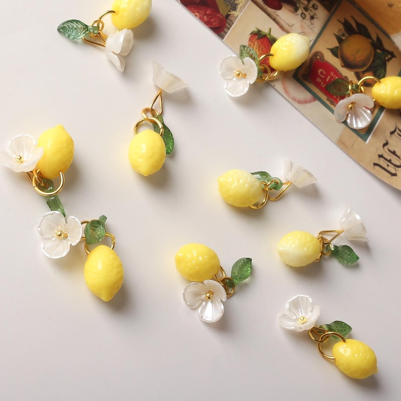 May include: A collection of lemon-themed jewelry, including earrings and charms. Each piece features a bright yellow lemon, delicate white flowers, and green leaves, all connected with gold-colored metal. The earrings and charms are arranged on a white surface.