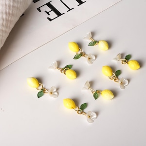 May include: Lemon-shaped earrings with gold-colored accents, green leaves, and white flower details. The earrings are arranged on a white surface, creating a fresh and summery aesthetic. The earrings are a charming accessory.