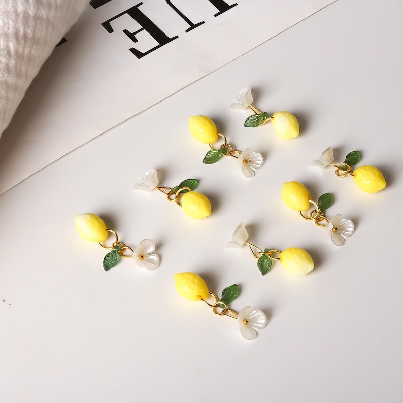 May include: Lemon-shaped earrings with gold-colored accents, green leaves, and white flower details. The earrings are arranged on a white surface, creating a fresh and summery aesthetic. The earrings are a charming accessory.
