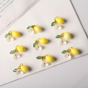 May include: A collection of yellow lemon charms with gold-colored loops, green leaves, and white flower accents. The charms are arranged on a white surface, creating a bright and cheerful aesthetic. These charms are ideal for jewelry making or craft projects.