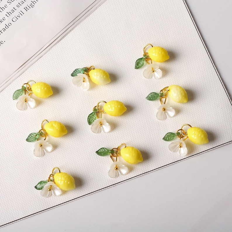May include: A collection of yellow lemon charms with gold-colored loops, green leaves, and white flower accents. The charms are arranged on a white surface, creating a bright and cheerful aesthetic. These charms are ideal for jewelry making or craft projects.