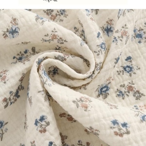 Pode incluir: Um tecido de cor creme com uma superfície texturizada e enrugada, apresentando um padrão repetitivo de pequenos grupos de flores azuis e castanhas. O texto YO CRAFT ART está impresso no topo da imagem acima do tecido, acompanhado por uma pequena ilustração botânica.