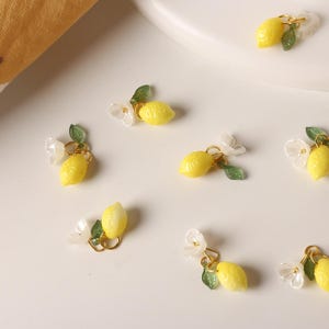 May include: Close-up of several small, yellow lemon charms with gold-colored hoops, green leaves, and white flower accents. The charms are scattered on a white surface, suggesting a jewelry or craft theme.