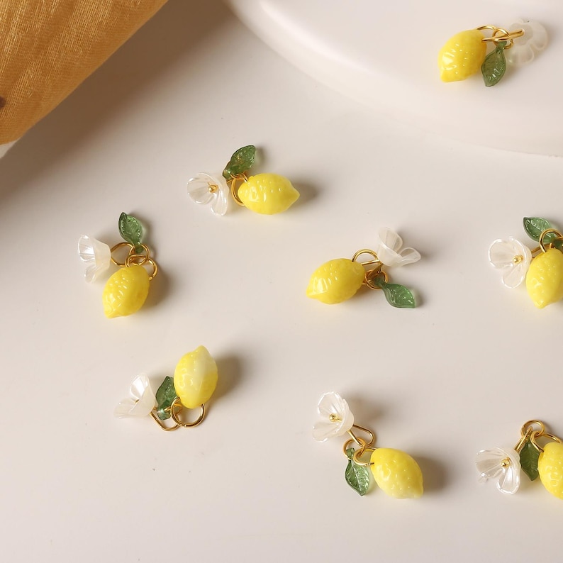 May include: Close-up of several small, yellow lemon charms with gold-colored hoops, green leaves, and white flower accents. The charms are scattered on a white surface, suggesting a jewelry or craft theme.