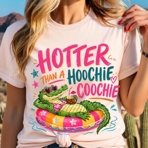 May include: Light pink t-shirt with the text "HOTTER THAN A HOOCHIE COOCHIE" in pink and teal. A cartoon alligator wearing sunglasses and a bikini floats on a colorful inflatable ring, holding a tropical drink.