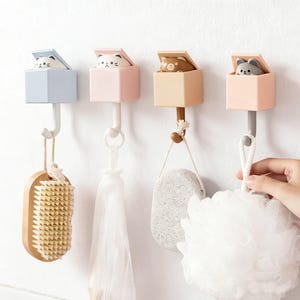 May include: Four decorative wall hooks with animal-themed designs. Each hook features a small box with a different animal figure peeking out. The hooks are in pastel colors and hold various bathroom accessories, including a brush and a loofah.