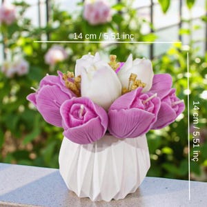 May include: A floral arrangement of purple and white tulip-shaped candles in a white geometric vase. The arrangement is approximately 14 cm (5.51 inches) tall and wide, set against a blurred green backdrop.