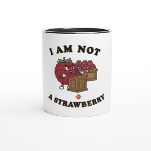 Peut inclure: Mug en céramique blanche avec un bord noir. Il présente un dessin animé de framboise à un pupitre, avec le texte "I AM NOT A STRAWBERRY". La framboise est rouge avec des feuilles vertes. La tasse a une finition brillante.