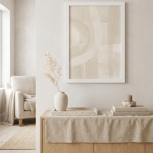 Puede incluir: Una lámina de arte abstracto enmarcada con formas circulares en tonos beige y crema, expuesta sobre un aparador de madera clara. El aparador sostiene un jarrón beige texturizado con tallos secos, una bandeja de piedra y libros apilados.