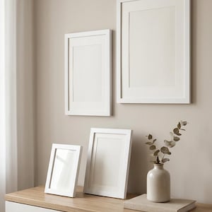Puede incluir: Dos marcos de fotos rectangulares blancos montados en una pared, con otros dos marcos blancos sobre una superficie de madera junto a un jarrón de cerámica con una rama de eucalipto seco que descansa sobre un libro beige.
