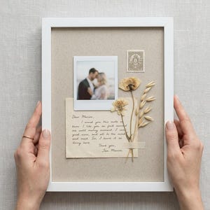 Könnte beinhalten: Ein weißer rechteckiger Rahmen mit beigem Passepartout, einem quadratischen Foto eines Paares, einer alten Briefmarke, einem handschriftlichen Brief an Marion sowie getrockneten Pflanzen, darunter eine Blume und Getreidehalme.