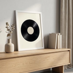 May include: A framed wall art print featuring a centred black vinyl record with a cream-coloured centre label. The artwork is encased in a simple white frame, showcasing a minimalist composition that highlights the circular shape of the record.