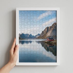 May include: A jigsaw puzzle in a white frame featuring a landscape photograph of a red cabin on a rocky shoreline by a calm body of water. In the background, tall, jagged mountains rise under a blue sky with light clouds, creating a reflection.