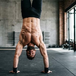 May include: A person doing a handstand in a gym setting. They are wearing black shorts and wrist supports. The background includes a concrete wall, a window, and gym equipment.