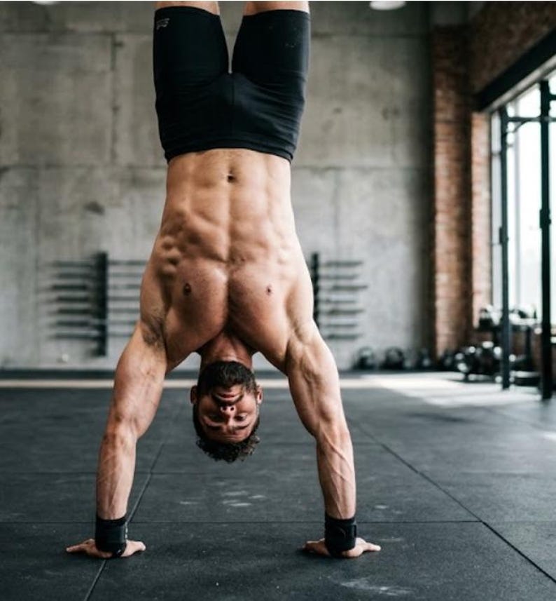 May include: A person doing a handstand in a gym setting. They are wearing black shorts and wrist supports. The background includes a concrete wall, a window, and gym equipment.
