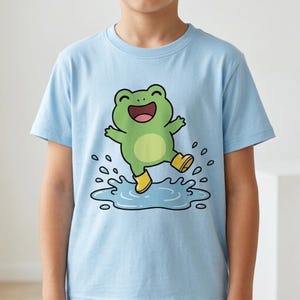 Op de afbeelding: Een lichtblauw T-shirt met korte mouwen en een grafische illustratie van een groene kikker met gele regenlaarzen, die in een blauwe waterplas met spetters springt. De kikker heeft een vrolijke uitdrukking met een open mond en armen omhoog.