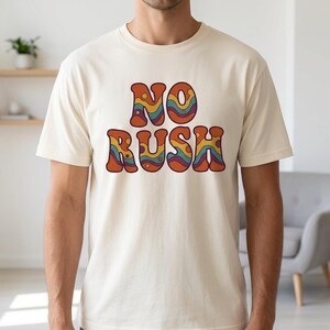 May include: A cream-coloured t-shirt featuring the words "NO RUSH" in a retro, wavy font. The text is in shades of orange, blue, and yellow. The shirt is short-sleeved and appears to be made of cotton.