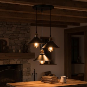 May include: Three-light pendant fixture with black metal shades and brass-coloured accents. The light fixtures hang from a black ceiling plate. The bulbs are exposed and emit a warm glow. The fixture is in a rustic-style room.