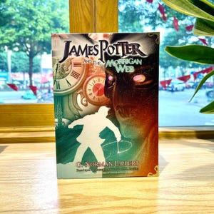 May include: A book titled James Potter and the Morrigan Web by G. Norman Lippert. The cover features a white silhouette of a person, two clock faces, and a dark, glowing face with bright eyes. The design uses shades of green, red, and black.