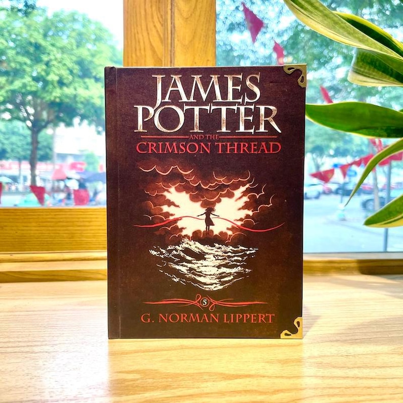 May include: A hardcover book titled James Potter and the Crimson Thread by G. Norman Lippert. The dark brown cover features a central illustration of a figure standing on waves beneath clouds, holding a red thread, with gold corner accents.