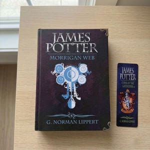 May include: A hardcover book titled James Potter and the Morrigan Web by G. Norman Lippert with a dark purple cover featuring a central clock graphic and metal corner protectors, accompanied by a matching vertical bookmark with a crest design.