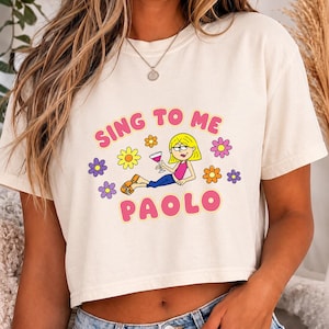 May include: A cream-colored short-sleeve t-shirt featuring a graphic of a cartoon character with blonde hair holding a cocktail glass, surrounded by colorful flowers. The text "SING TO ME" and "PAOLO" is printed in pink with a yellow outline.