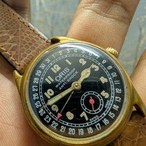 Puede incluir: Un reloj Oris vintage dorado con esfera negra y correa de cuero marr&oacute;n. La esfera muestra n&uacute;meros blancos, un indicador de fecha y las palabras "WATERPROOF ANTI-SHOCK". El reloj tiene agujas doradas y un segundero.