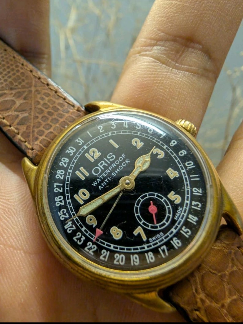 Puede incluir: Un reloj Oris vintage dorado con esfera negra y correa de cuero marr&oacute;n. La esfera muestra n&uacute;meros blancos, un indicador de fecha y las palabras "WATERPROOF ANTI-SHOCK". El reloj tiene agujas doradas y un segundero.