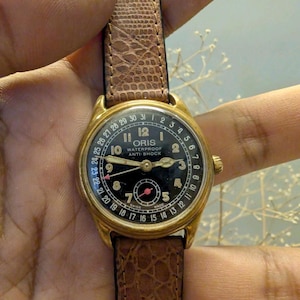 Puede incluir: Un reloj de pulsera Oris vintage dorado con esfera negra y correa de cuero marr&oacute;n. La esfera del reloj presenta n&uacute;meros y agujas blancas, con el nombre de la marca y el texto "WATERPROOF ANTI-SHOCK". La correa es de cuero marr&oacute;n texturizado.