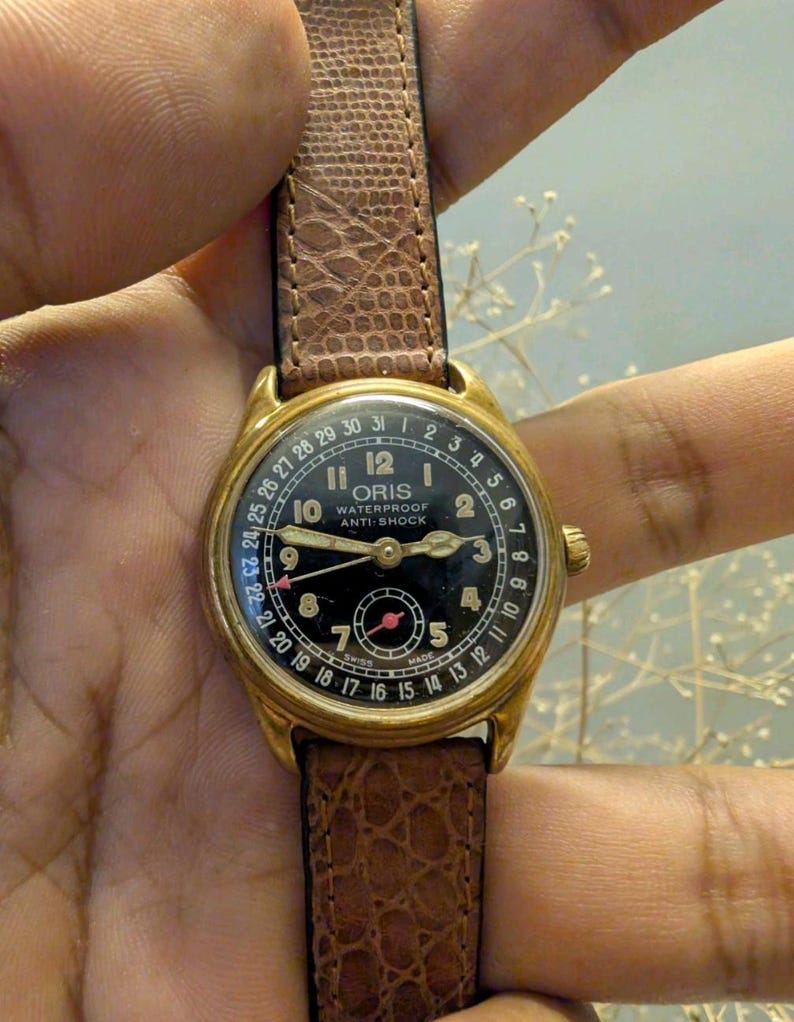 Puede incluir: Un reloj de pulsera Oris vintage dorado con esfera negra y correa de cuero marr&oacute;n. La esfera del reloj presenta n&uacute;meros y agujas blancas, con el nombre de la marca y el texto "WATERPROOF ANTI-SHOCK". La correa es de cuero marr&oacute;n texturizado.