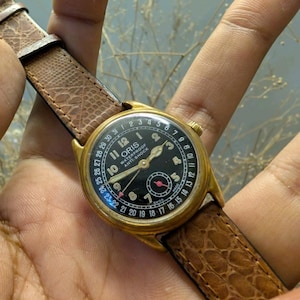Puede incluir: Un reloj de pulsera Oris vintage dorado con esfera negra y correa de cuero marrón. La esfera del reloj muestra números y manecillas blancas, con una pequeña esfera roja. La correa es texturizada y el reloj se sostiene en una mano.