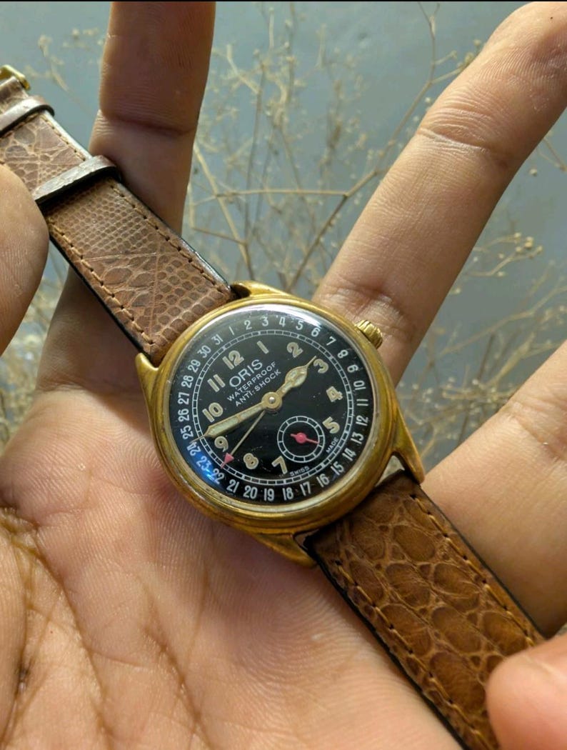 Puede incluir: Un reloj de pulsera Oris vintage dorado con esfera negra y correa de cuero marr&oacute;n. La esfera del reloj muestra n&uacute;meros y manecillas blancas, con una peque&ntilde;a esfera roja. La correa es texturizada y el reloj se sostiene en una mano.